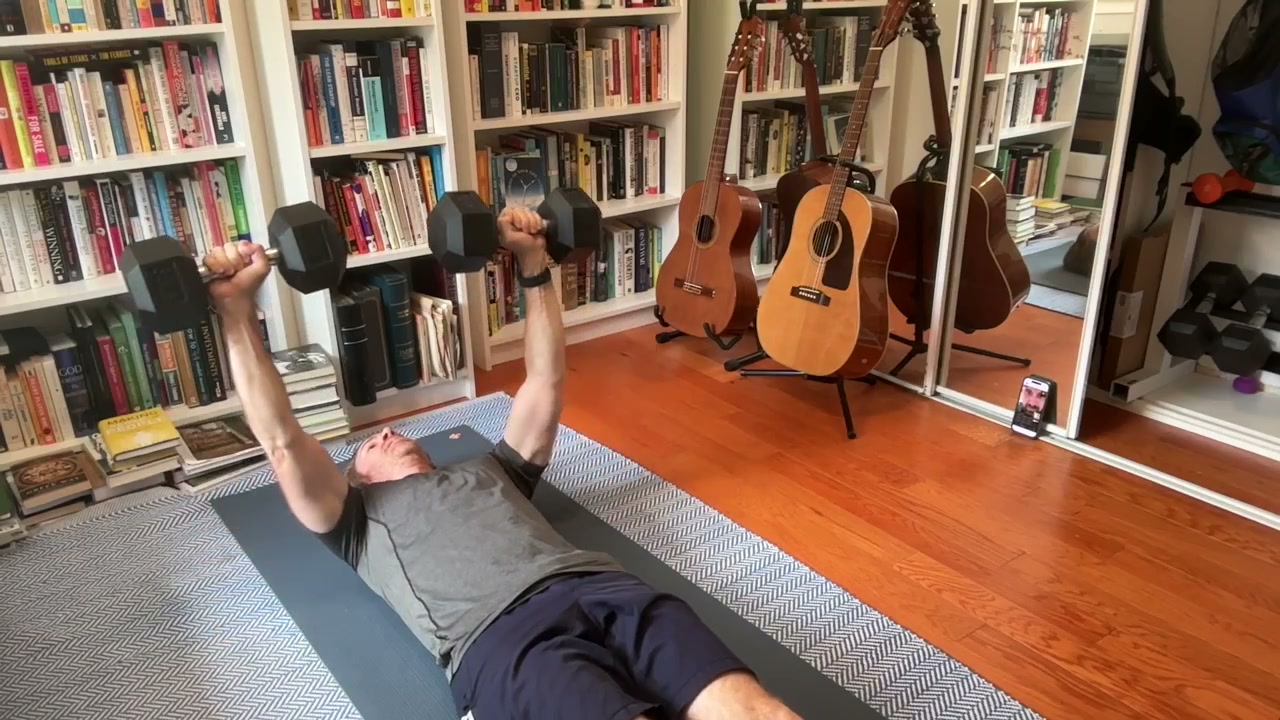 A client pressing dumbbells on a mat at home while his coach watches live from a propped-up phone.